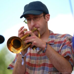 Man wearing a dark cap and glasses playing a brass trumpet outdoors, close up on the instrument and his lips touch the mouthpiece.