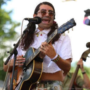 Musician with long hair and sunglasses singing into a microphone while playing an acoustic guitar on stage outdoor venue.