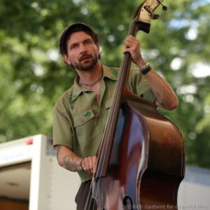 Musician playing a double bass outdoors, wearing a green shirt, hat, and bracelets, with visible tattoos on his arms.