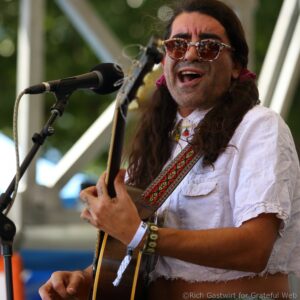 Male singer with long hair and patterned sunglasses singing into a microphone on stage, holding an acoustic guitar.