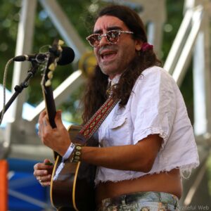 Male guitarist with long hair and floral glasses singing into a microphone on an outdoor stage while playing an acoustic guitar
