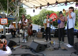 Outdoor festival band on stage: guitarist, saxophonist, trombonist, keyboardist perform as audience shoots from front row with phones; trees in background and a red festival sign nearby.