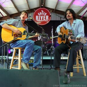 Two male musicians play acoustic guitars on a stage, singing into a microphone with a red Green River Festival sign behind them.