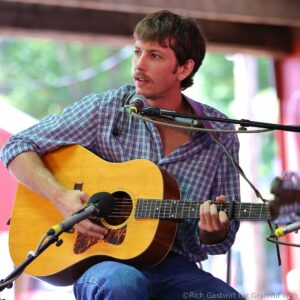 Male guitarist singing into a microphone while playing an acoustic guitar on stage.