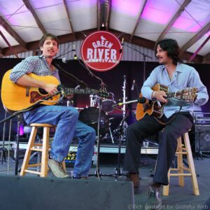 Two male musicians seated on stools playing acoustic guitars on a festival stage with a red 'Green River Festival' sign behind them.