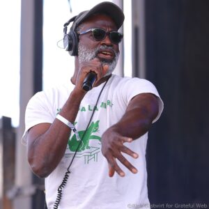 Male performer wearing a cap and sunglasses, holding a microphone and wearing over‑ear headphones on stage.