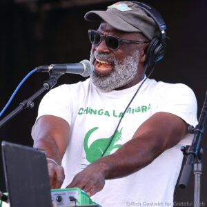 Male singer wearing a cap and sunglasses, singing into a microphone on stage with headphones on