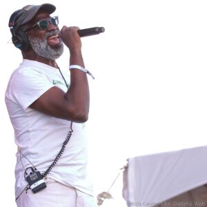 Man wearing a cap and sunglasses, singing into a handheld microphone with over-ear headphones on.