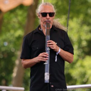 Man with long gray hair and sunglasses playing a wind instrument outdoors.