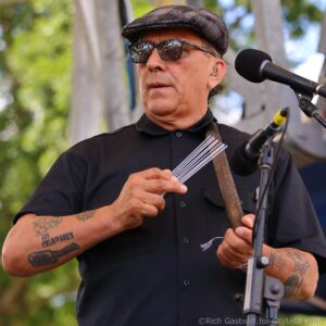 Male musician wearing a flat cap and sunglasses, singing into a microphone outdoors, with visible tattoos on his forearms.