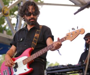 Musician with dark hair and sunglasses plays a red bass guitar on an outdoor stage.