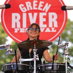 Drummer wearing sunglasses plays a drum kit in front of a large red circular sign that reads GREEN RIVER FESTIVAL at an outdoor event.