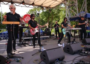 Outdoor concert stage with a five-member band playing guitars and keyboards under a canopy, trees in the background, several amplifiers and cables on the floor.