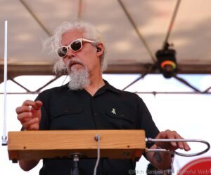 Bearded musician wearing white sunglasses plays a wooden keyboard on a stage under a tent.