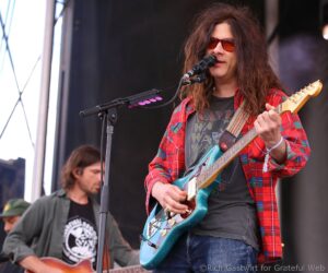 Musician with long hair and red sunglasses plays a turquoise electric guitar on stage, wearing a red plaid shirt.