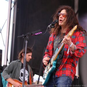 Male guitarist in red plaid shirt singing into a microphone on stage with a teal electric guitar, sunglasses on, sunlit backdrop