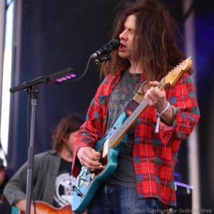Guitarist singing into a microphone on stage, wearing a red plaid shirt and playing a blue electric guitar.