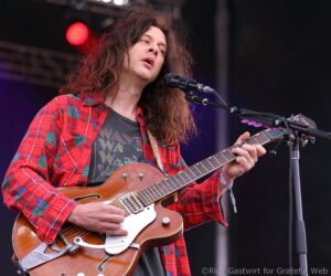 Musician with long hair and red plaid shirt singing into a microphone while playing a sunburst electric guitar on stage.