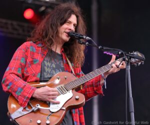 Guitarist with long wavy hair in a red plaid shirt singing into a microphone while playing a sunburst electric guitar on stage on a live performance.