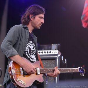 Guitarist playing a sunburst electric guitar on stage with purple lighting and ear monitors in his ear.