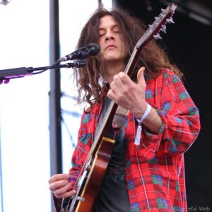Musician with long hair in a red plaid shirt, playing an electric guitar and singing into a microphone on stage.