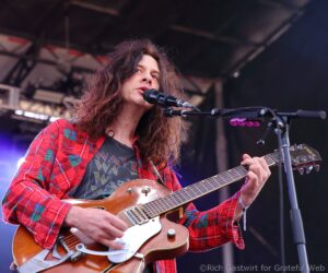 Male singer with long hair in a red plaid shirt, playing a sunburst electric guitar and singing into a microphone on stage behind a pedalboard.