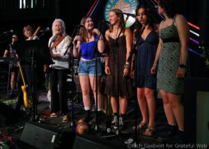 A group of women singing on a stage with multiple microphones, guitars, and a drummer visible in the background during a live performance.