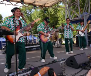 Four musicians in matching green-and-white geometric shirts perform on an outdoor stage, each playing electric guitars and singing into microphones.