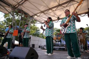 Five musicians in matching green-and-white patterned shirts perform on an outdoor stage with guitars and drums.",