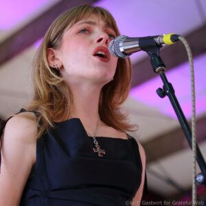 Woman singing into a silver microphone on stage with purple lighting in the background, wearing a dark dress and silver necklace.