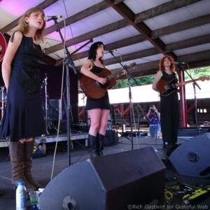 Three female musicians perform on an outdoor stage: two with acoustic guitars singing into microphones, one foreground guitarist in a black outfit and tall boots.