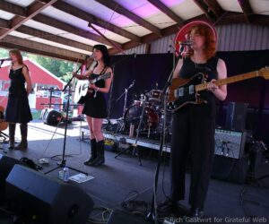 Three female musicians performing on an outdoor covered stage with microphones and guitars.
