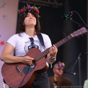 Woman wearing a colorful flower crown plays an acoustic guitar on stage with a microphone nearby.