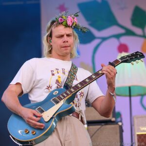 Male guitarist wearing a flower crown and white t-shirt, playing a teal electric guitar on stage with a colorful backdrop on stage lighting behind him.