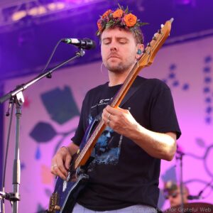 Male musician wearing a flower crown while playing a bass guitar on stage, singing into a microphone with purple stage lighting in the background.