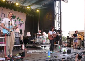 Band performing live on an outdoor stage; foreground guitarist wearing a flower crown sings into a microphone while others play instruments in the background.