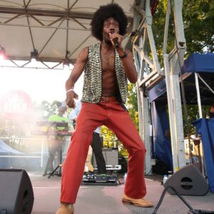 Male singer with an afro on stage, holding a microphone and wearing a patterned sleeveless vest and red pants during a festival performance.
