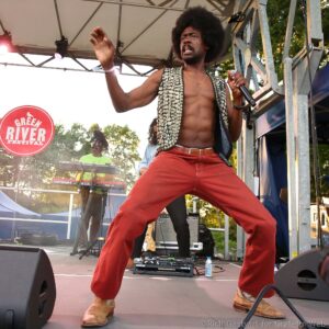 Male performer with an afro singing into a microphone on an outdoor stage, wearing a studded sleeveless vest and red pants.