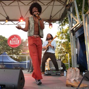 Male singer with an afro, wearing a patterned sleeveless vest and red pants, singing into a microphone on an outdoor festival stage with a guitarist in the background and a Green River Festival sign visible.