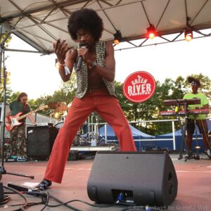 Male performer with an afro singing into a microphone on an outdoor stage, wearing a studded vest and red pants.