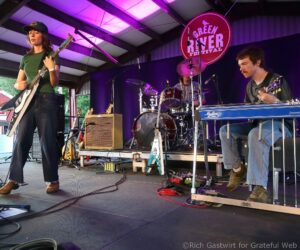 Band performing on stage at Green River Festival; left guitarist in green shirt and cap, center drummer, right keyboardist with pedal steel guitar under purple lights.