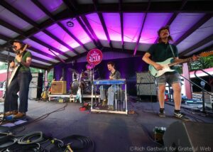 Band performing on an outdoor stage with purple lighting; left guitarist sings into a microphone, right guitarist plays a teal Stratocaster, drummer in back, amps and cables visible amid instruments and gear.