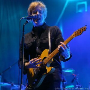 Guitarist singing into a microphone on blue-lit stage while playing a yellow electric guitar