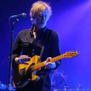 Male guitarist performing on stage, blue lighting, playing a yellow electric guitar near a microphone stand.