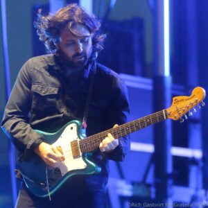 Male guitarist with a beard and long hair, playing a teal electric guitar on a stage bathed in blue light.