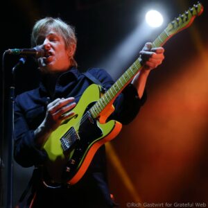 Male guitarist sings into a microphone while playing a bright lime-yellow electric guitar on stage under lights