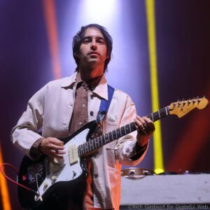 Guitarist performing on stage, playing a black electric guitar under purple lighting and yellow vertical light bars.