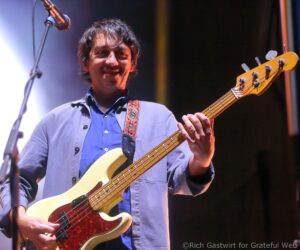 Smiling male guitarist on stage playing a cream Telecaster-style electric guitar with a floral strap.