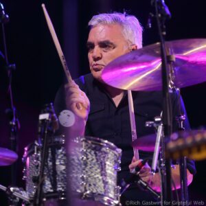 Close-up of a male drummer with gray hair playing drums on stage under purple lights making focused expression.