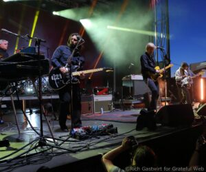 Live rock band on stage with a center guitarist, other musicians and gear visible, colorful stage lights and cables on the floor.